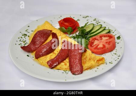 Omelette à la saucisse délicieuse dans l'assiette, à côté du concombre de tomates et du persil, isolée sur fond blanc Banque D'Images