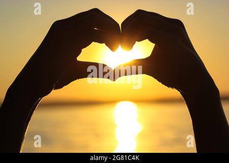 Silhouette des mains de la femme en forme de coeur au coucher du soleil contre un ciel jaune-orange, des rayons de soleil et un soleil à l'intérieur. Symbole de l'amour. Vacances par a s Banque D'Images