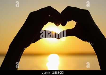 Silhouette des mains de la femme en forme de coeur au coucher du soleil contre un ciel jaune-orange, des rayons de soleil et un soleil à l'intérieur. Symbole de l'amour. Vacances par a s Banque D'Images