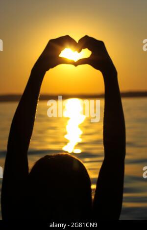 Silhouette des mains de la femme en forme de coeur au coucher du soleil contre un ciel jaune-orange, des rayons de soleil et un soleil à l'intérieur. Symbole de l'amour. Vacances par a s Banque D'Images