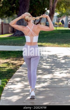 Femme blonde atractive en bonne santé et en bonne forme qui s'adapte à sa longue queue de cheval tout en marchant dans le parc. Banque D'Images