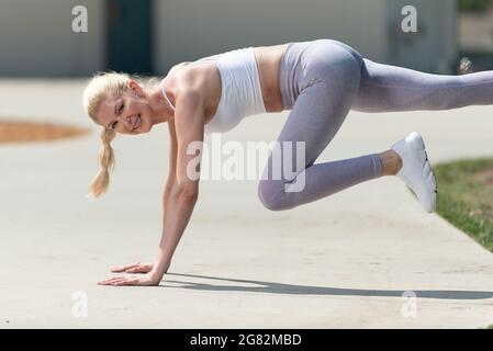 Une femme blonde atractive en bonne santé et en forme, qui fait un pont sur le trottoir du parc pour renforcer son cœur tout en souriant avec enthousiasme. Banque D'Images