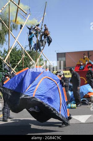 Scunthorpe, Royaume-Uni. 16 juillet 2021. Les policiers délèvent des tentes de protestation tandis que les manifestants rebelles d'animaux regardent avec des torches de fumée au sommet d'une structure en bambou pendant la manifestation. Plus de 50 manifestants de la rébellion animale ont fermé la seule usine de distribution de hamburgers de McDonald's, l'usine OSI Food Solutions au Royaume-Uni, en blockant l'entrée avec des balises en bambou et des véhicules. Les rebelles exigent que McDonald's passe à des menus entièrement basés sur des plantes d'ici 2025 et mette fin à la souffrance de milliards de vies et pour éviter la crise climatique tout en sauvant la forêt amazonienne. Crédit : SOPA Images Limited/Alamy Live News Banque D'Images