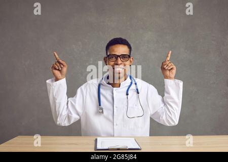 Un médecin afro-américain souriant assis sur un bureau, pointant vers le haut Banque D'Images