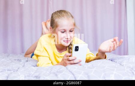Une adolescente de race blanche avec un smartphone dans ses mains, allongé sur le lit, blogging, communiquant sur les réseaux sociaux avec ses amis, dépensant gratuitement Banque D'Images