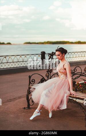 une ballerine fragile dans une longue robe rose avec une jupe organza complète se trouve sur un banc en bois de fer forgé sur la rive de la rivière dans un paysage urbain Banque D'Images