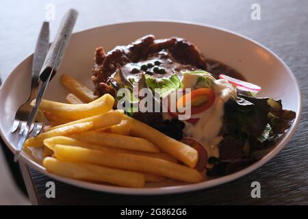 Steak de poulet fraîchement cuit avec sauce au poivre, frites et salade, servi sur une grande assiette en céramique avec des ustensiles en métal Banque D'Images