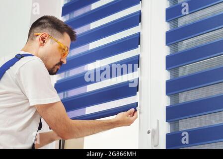 Menuisier de construction mâle tirant vers le bas un bleu jalousie sur une fenêtre. Réparation et inspection des rideaux en tissu. Système Duo jour et nuit aveugle. Élément de décoration intérieure. Banque D'Images