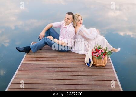 Un couple mûr et paisible se reposant dos à dos sur un pont en bois au bord de la rivière ou de l'étang, en profitant de la vue sur la nature et le paysage Banque D'Images