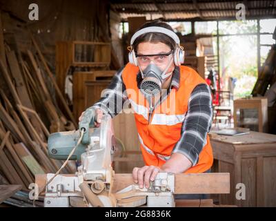 Beau artisan de charpentier caucasien portant un équipement de sécurité travaillant avec du bois dur à l'aide d'une scie à onglets pour couper un morceau de bois dans l'atelier. Fabrication Banque D'Images