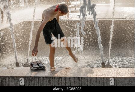 Garçon lavant son pied dans des jets de fontaine d'eau, vu à travers des pulvérisations et des gouttelettes, en été pendant la vague de chaleur Banque D'Images