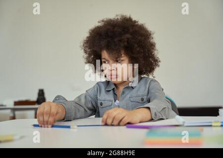 Un petit écolier mécontent avec des cheveux afro qui ont l'air triste en étant assis à la table dans la salle de classe de l'école primaire Banque D'Images