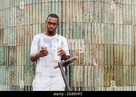 homme de banlieue africain debout dans la rue près d'un scooter et utilisant son téléphone Banque D'Images