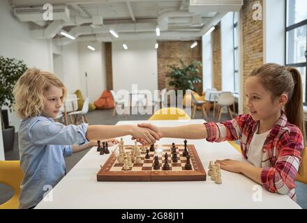 Respect. Adorables petits amis, un garçon caucasien et une fille se serrant la main après le match, jouant aux échecs, assis ensemble à la table à l'école Banque D'Images