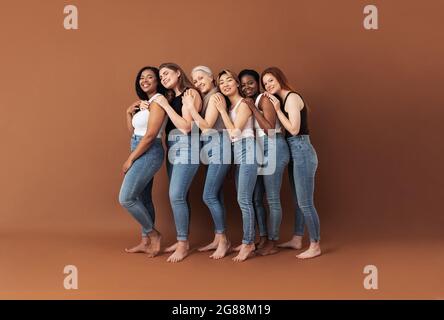 Pleine longueur de six femmes souriantes embrassant en studio. Les femelles de différents âges et types de corps posant sur fond brun. Banque D'Images
