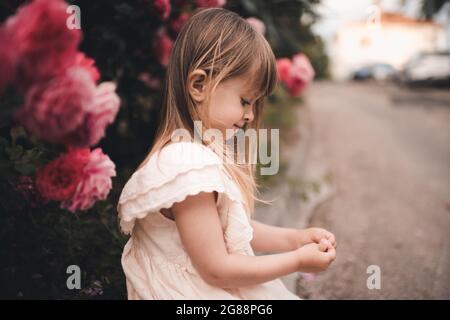 Drôle mignon enfant fille 3-4 ans vêtements élégant posant avec fleurs roses roses dans le parc à la rue de la ville à l'extérieur de près. Saison d'été. CH Banque D'Images