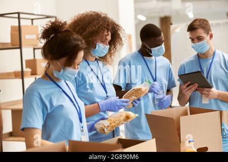 Une jeune équipe de bénévoles dans le tri des masques et des gants de protection, l'emballage des aliments dans des boîtes en carton, travaillant sur le projet de don à l'intérieur Banque D'Images