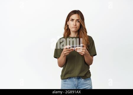 Contrarié femme adulte tenant le smartphone dans les deux mains, déçu par le jeu vidéo ou l'application, froning et smirking insatisfait, se tenant contre blanc Banque D'Images