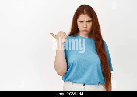 Une jeune fille à tête rouge en colère et grognante sillonnait les sourcils, pointant le pouce vers la gauche et se frognant détestait, se plaignant, se lassant du smth injuste, se tenant debout en chemise Banque D'Images