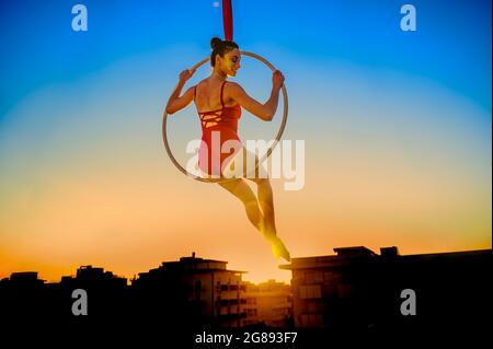 Acrobat aérien dans un cerceau au coucher du soleil sur la ville d'horizon - activité en plein air de gymnaste se présentant sur le cerceau dans le ciel - Girl interprète le e acrobatique Banque D'Images