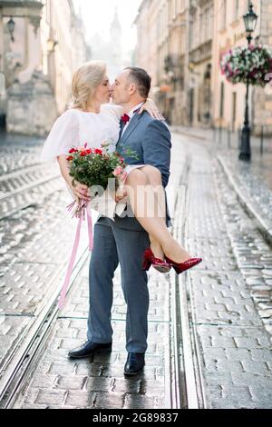 Charmant couple mûr en plein air dans la vieille ville souriant et regardant l'un l'autre. Temps ensemble. Le beau homme tient sa femme sur les mains Banque D'Images