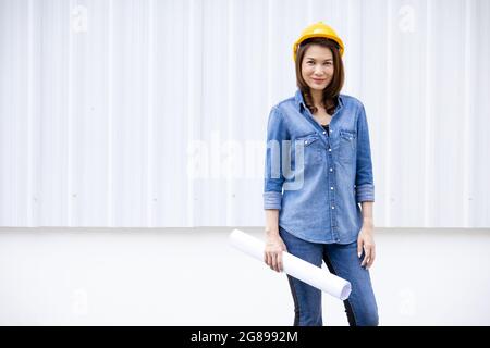 Belle asiatique femme en Jean robe portant un chapeau de sécurité jaune et tenir le rouleau de papier blanc debout devant de la paroi en tôle à la construction Banque D'Images