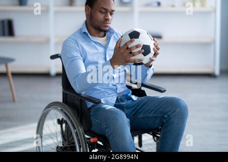 Homme noir handicapé avec ballon de football assis en fauteuil roulant, se sentant déprimé au-dessus de sa blessure à la maison, foyer sélectif Banque D'Images