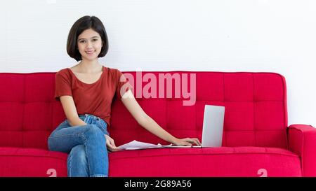 Jeune et beau cheveux courts moitié thaï - fille allemande assis sur un canapé rouge et utilisant un ordinateur portable pour travailler de la maison avec un visage souriant et happi Banque D'Images