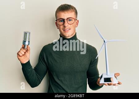 Jeune homme caucasien tenant un moulin solaire et une ampoule LED souriant avec un sourire heureux et frais sur le visage. Montrant des dents. Banque D'Images