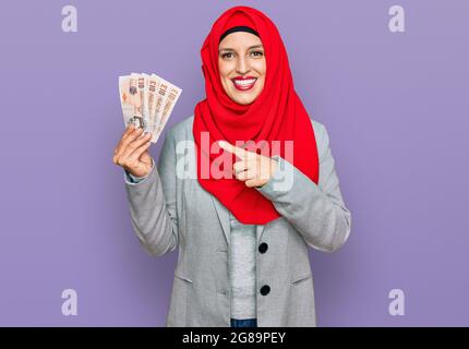 Belle femme hispanique portant le hijab islamique tenant 10 livres de billets souriant souriant souriant pointant avec la main et le doigt Banque D'Images