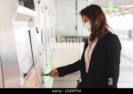 Belle femme asiatique dans un costume noir portant un masque de protection d'hygiène médicale achetant métro ou train de ciel forme distributeur automatique à la gare. Concept pour Banque D'Images