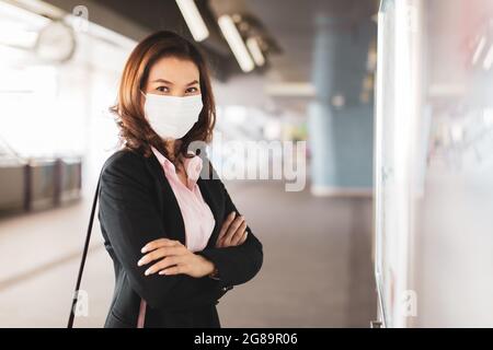 Belle femme asiatique dans un costume noir portant un masque de protection d'hygiène médicale debout au tableau de bord de la gare, regardant la caméra. Concept pour W Banque D'Images