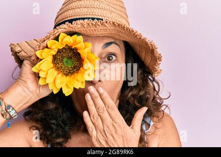 Femme hispanique d'âge moyen portant un chapeau d'été tenant le tournesol sur l'oeil couvrant la bouche avec la main, choquée et peur de l'erreur. Expression surprise Banque D'Images