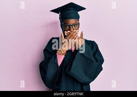 Une jeune fille afro-américaine portant une casquette de remise des diplômes et une robe de cérémonie a été choquée en couvrant la bouche avec des mains pour erreur. Concept secret. Banque D'Images