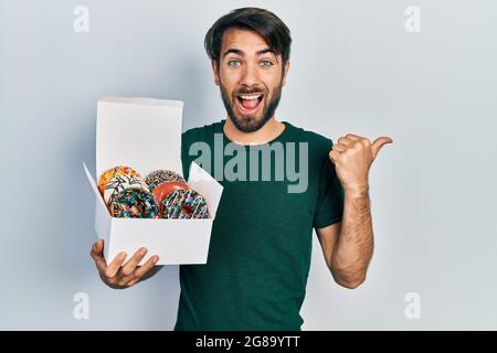 Jeune homme hispanique tenant une boîte de beignets colorés savoureux pointant le pouce vers le côté souriant heureux avec la bouche ouverte Banque D'Images