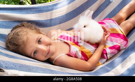 Un lapin blanc moelleux dans les bras d'une jolie fille. L'enfant tient un lapin décoratif dans ses bras et roule sur un hamac par temps ensoleillé. Bon petit g Banque D'Images