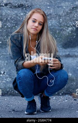 Jeune femme blonde tendance qui s'accroupite pour écouter sa musique sur un téléphone portable et des écouteurs qui regardent attentivement l'appareil photo avec un sourire tranquille Banque D'Images