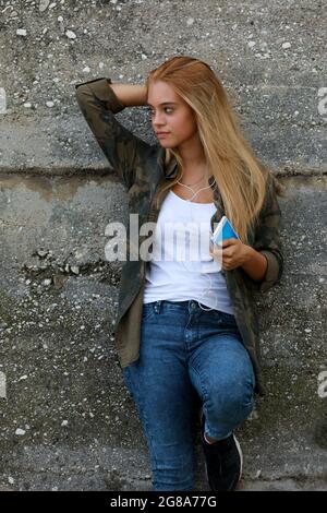 Jeune femme blonde moderne à la mode qui écoute de la musique sur son mobile alors qu'elle se détend contre un mur de béton abîmé en regardant de côté avec un pensif Banque D'Images