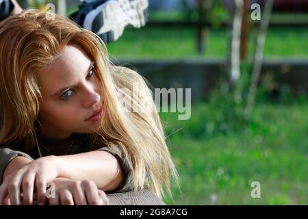 Belle jeune femme blonde regardant avec attention la caméra pendant qu'elle se détend sur un banc en bois dans le parc dans un gros plan partiel bas angle Banque D'Images