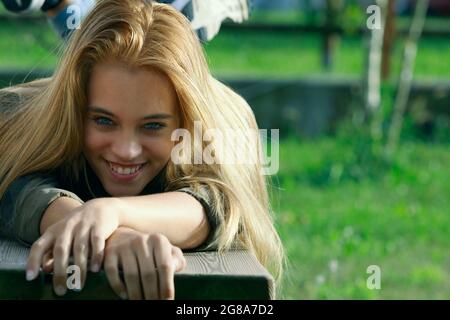 Jolie jeune femme blonde avec un sourire vivace couché sur son ventre sur un banc de jardin dans une vue frontale à angle bas avec copyspace Banque D'Images