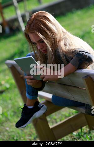 Jeune femme blonde se détendant drapée sur un banc de bois dehors dans le jardin au coucher du soleil pour lire ou regarder les médias sur une tablette Banque D'Images