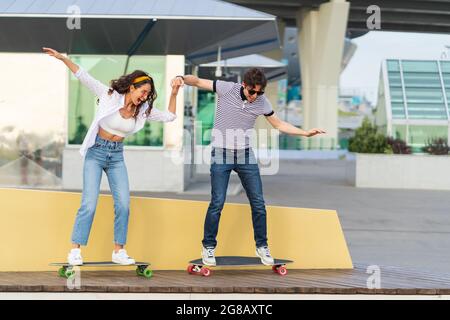 Un couple de jeunes patineurs actifs apprend à monter en planche à roulettes ensemble. Tenez les mains riant sur le stand du skateboard Banque D'Images