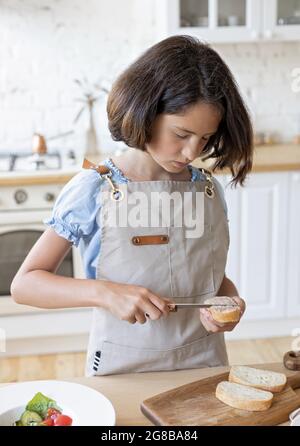 Une petite brune fait des sandwichs dans la cuisine. Belle fille cuisine dans la cuisine. Belle fille en tablier sur une maison de couleur claire Banque D'Images