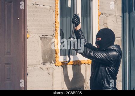 Cambrioleur avec visage obscurci essayant de briser la fenêtre. Un homme masqué brise une fenêtre avec un pied de biche. Vol de garage. Vol d'une maison privée. Lit bébé Banque D'Images