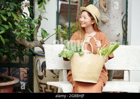 Portrait d'une jolie jeune femme positive assise sur un banc avec un sac d'épicerie fraîche qu'elle a acheté sur le marché local Banque D'Images