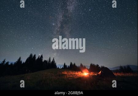 Campez de nuit sous un ciel étoilé et une voie lactée. Vue grand angle sur les paysages en montagne. Deux tentes et feu de camp, mur de spruces en arrière-plan. Concept de voyage et d'astrophotographie Banque D'Images