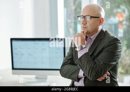 Homme d'affaires mature en lunettes et en costume debout au bureau et pensant à quelque chose Banque D'Images