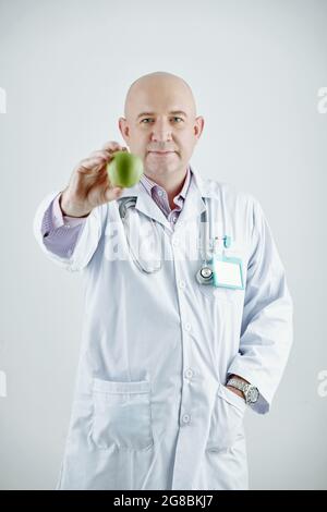 Portrait d'un médecin mature en manteau blanc tenant la pomme verte dans sa main et regardant l'appareil photo contre le fond blanc Banque D'Images