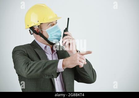 Ingénieur dans le casque de travail et dans le masque de protection pointant vers quelque chose tout en donnant des ordres sur la radio isolée sur fond blanc Banque D'Images