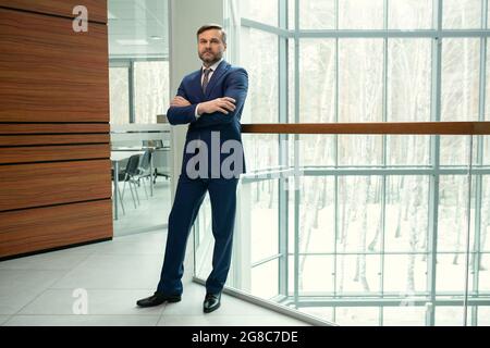 Portrait d'un homme d'affaires mûr et confiant en costume élégant debout avec ses bras croisés et regardant l'appareil photo tout en se tenant dans le hall de bureau Banque D'Images